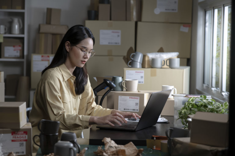 Woman working on laptop in e-commerce business setting with shipping boxes and plants